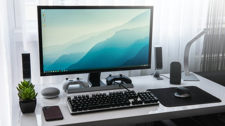 A computer monitor, keyboard, and game controllers on a white desk in a workspace
