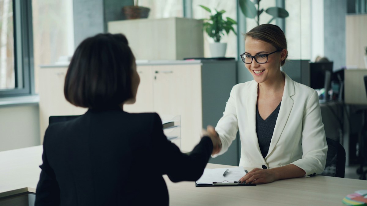 Two women shaking hands across an office desk, an illustrative interview photograph